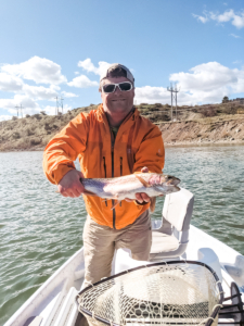 Dan wearing a bright orange jacket and sunglasses smiles at the camera as he proudly holds a trout out in front of him with both hands.
