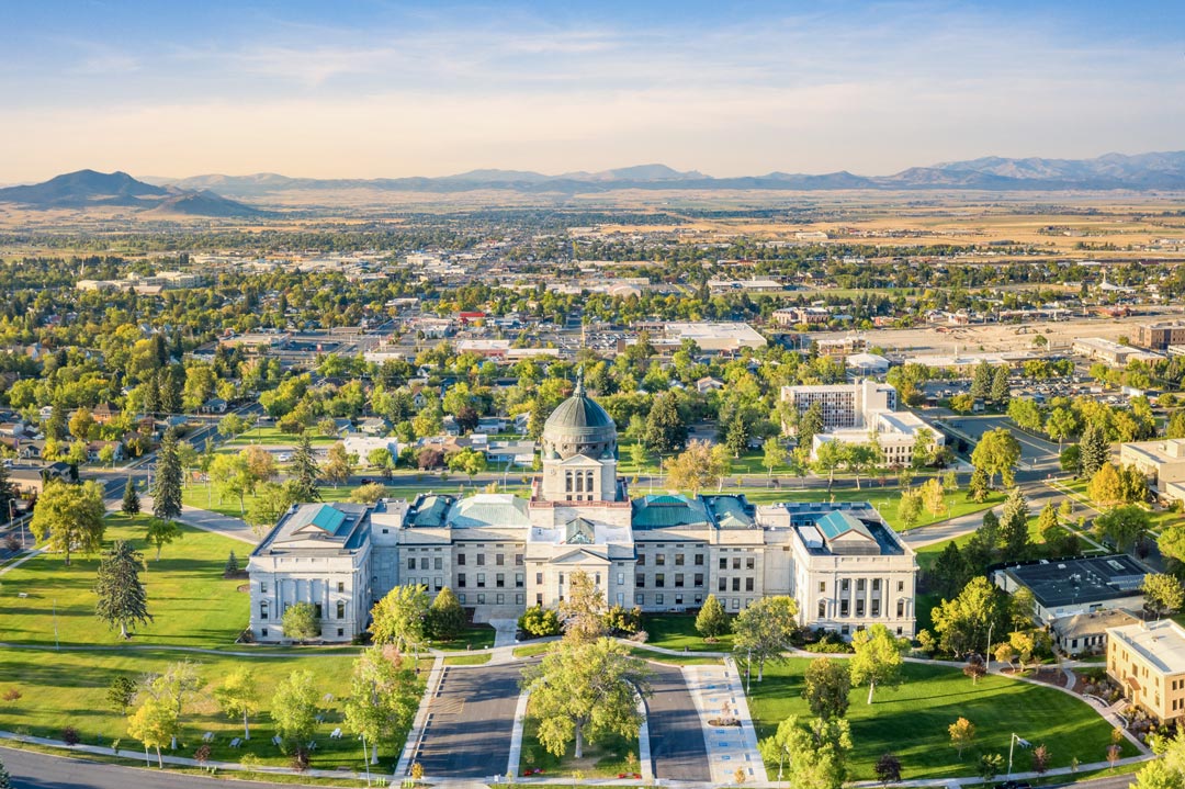 A drone photo in spring with a focus on the capital building in Helena, MT. The surrounding area fades to the mountains in the background.