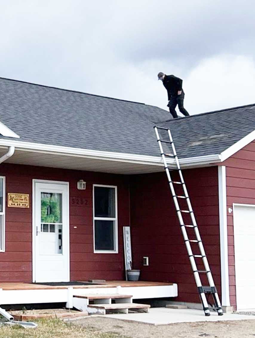 Roof Inspections All Mountain Roofing inquiries start with a roof inspection like the one depicted here. A ladder is carefully and correctly leaned against the red house with a white door. The roof being inspected is dark grey with shingles visibly missing to the right of the ladder over the garage. A person is on the roof slightly bent as they inspect the wear of the shingles looking for additional damage.