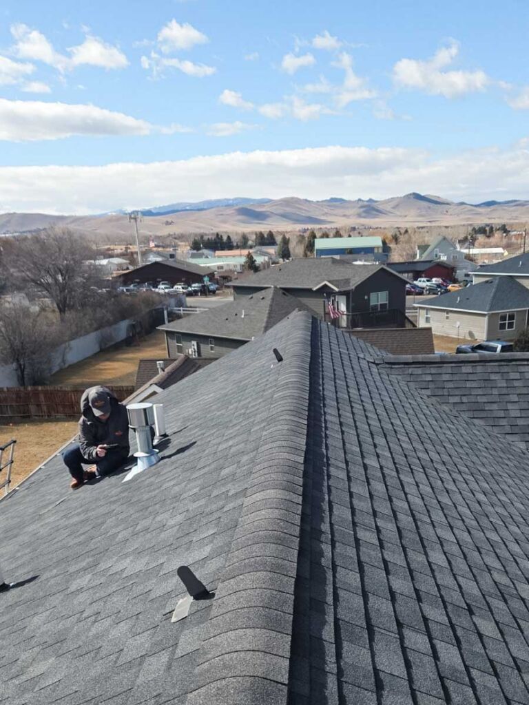 The Mountain Roofing team takes care to capture all of the details during roof inspections. One of our inspectors is kneeling on the roof to take a detailed note about the pipe flashing, the Helena, Montana landscape stretches out behind him with the roof line.